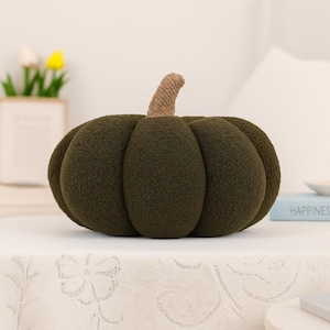 May include: A plush, olive green pumpkin-shaped pillow with a textured surface. The pillow has a light brown stem and is sitting on a white tablecloth. The pillow is a decorative item.