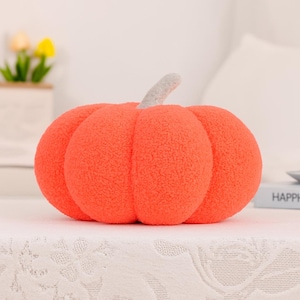 May include: A plush orange pumpkin with a textured surface and a gray stem. The pumpkin is a decorative item, perfect for fall or Halloween. It is sitting on a white surface.