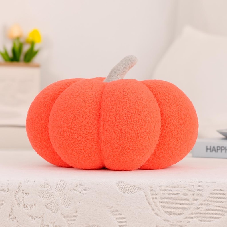 May include: A plush orange pumpkin with a textured surface and a gray stem. The pumpkin is a decorative item, perfect for fall or Halloween. It is sitting on a white surface.