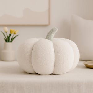 May include: A plush, white pumpkin-shaped pillow with a light green stem. The pillow is made of a soft, textured material and sits on a neutral-colored surface. The design is reminiscent of autumn decor.