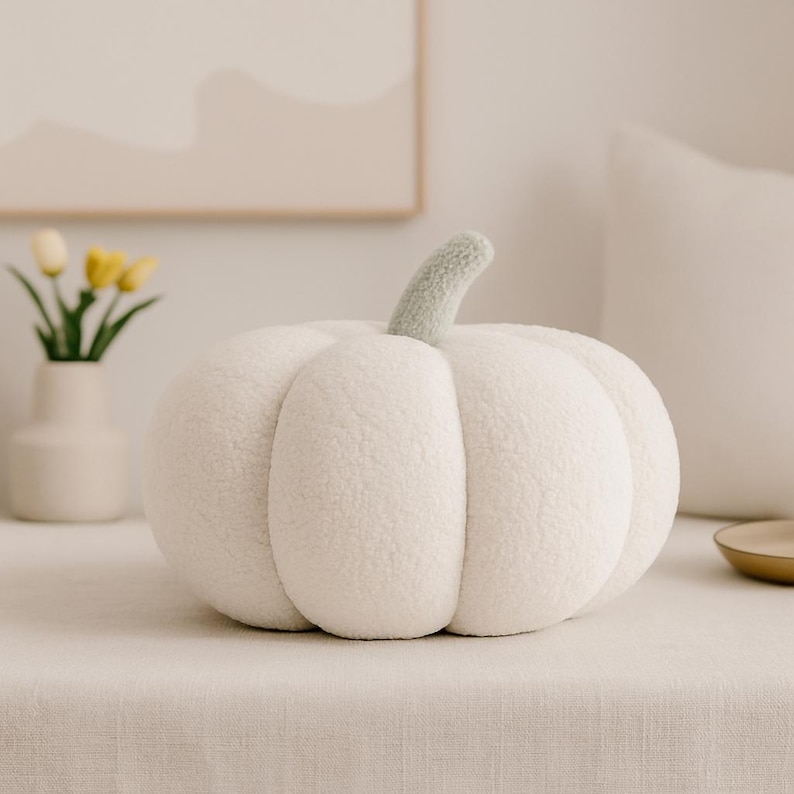 May include: A plush, white pumpkin-shaped pillow with a light green stem. The pillow is made of a soft, textured material and sits on a neutral-colored surface. The design is reminiscent of autumn decor.