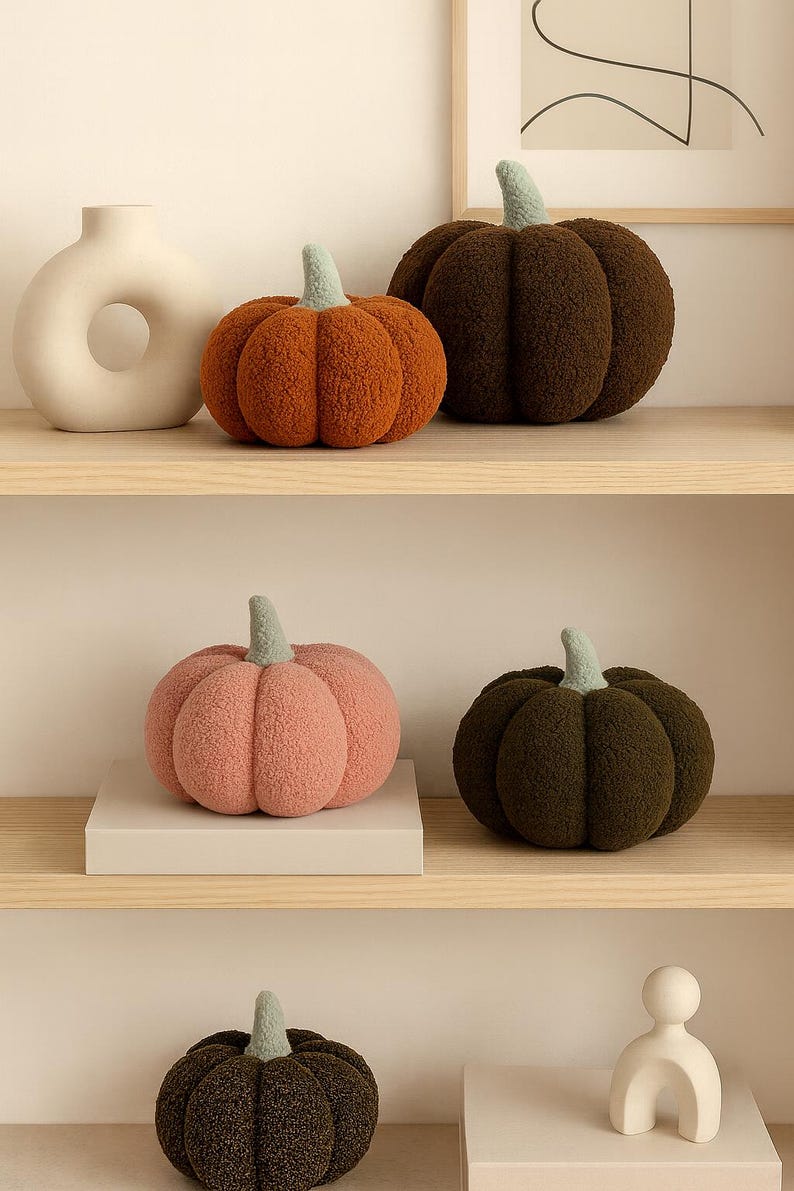 May include: A collection of plush pumpkin decorations in various colors, including orange, brown, pink, and green. Each pumpkin has a light green stem. The pumpkins are displayed on wooden shelves, alongside a white vase and a decorative sculpture.