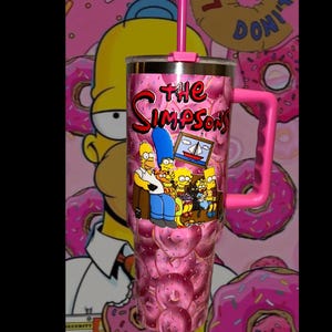 May include: A pink tumbler with a donut pattern and the text "The Simpsons." It has a pink handle and straw. The front shows the Simpson family. The background is pink with donut graphics. The tumbler is designed for cold beverages.