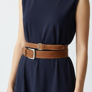 May include: A navy blue sleeveless dress with two brown leather belts, each with a silver buckle. The belts are a lighter shade of brown. The dress is a solid colour.