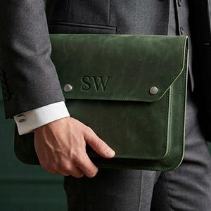 Op de afbeelding: Een donkergroene leren laptoptas met de initialen "SW" in reliëf op de voorkant. De tas heeft een klepsluiting met twee zilverkleurige drukkers. De tas wordt vastgehouden door een persoon in een grijs pak.