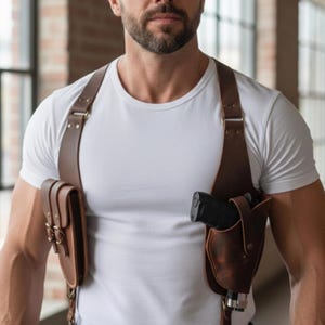 May include: A brown leather shoulder holster with two pouches. The holster has adjustable straps and metal buckles. One pouch holds a black handgun. The leather has a distressed finish.