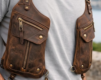 May include: A brown leather chest harness bag featuring two symmetrical compartments with brass-toned zippers, metal snap buttons, and adjustable straps. The design includes multiple zippered pockets and metal D-rings for secure storage and attachment.