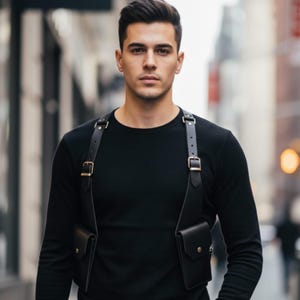 May include: A black leather chest harness featuring two symmetrical pockets with metallic snap closures, adjustable shoulder straps with silver-toned buckles, and a solid black long-sleeved shirt worn underneath the harness structure.