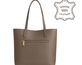 May include: A taupe-colored leather tote bag with a gold zipper closure and matching handles. The bag has a pebbled texture and a rectangular shape. The upper right corner features a brown graphic that reads "GENUINE 100% LEATHER".