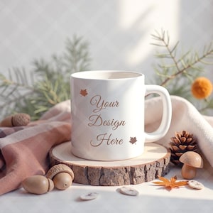 Könnte beinhalten: Eine weiße Keramiktasse mit dem Text "Your Design Here" in dekorativer Schrift, verziert mit Blätterillustrationen. Die Tasse steht auf einer Holzscheibe, umgeben von herbstlichen Dekorationen wie Eicheln, einem Tannenzapfen und einem Pilz.