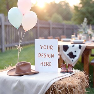 Op de afbeelding: Een rustieke buitenscène met een hooibaal tafel. Een bord met de tekst "YOUR DESIGN HERE" staat naast een bruine cowboyhoed en kleine bruine cowboylaarzen. Pastelkleurige ballonnen zweven erboven.