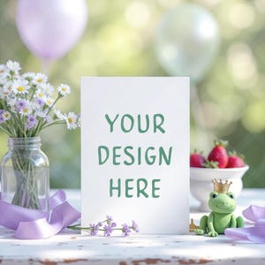 May include: A white card with the text "YOUR DESIGN HERE" in green, set on a white table. The scene includes a vase of daisies, strawberries, a frog figurine with a crown, and purple accents, with balloons in the background.