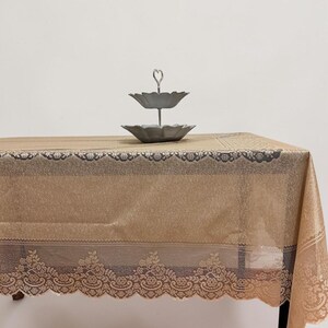 Puede incluir: Un mantel beige con un borde de encaje decorativo y una bandeja de dos niveles. El mantel está sobre una mesa y la bandeja es de metal.