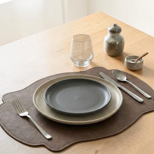 May include: A dark brown scalloped leather placemat holding a large speckled beige dinner plate topped with a smaller matte charcoal gray plate. A stainless steel fork, knife, and spoon are arranged alongside a clear glass and a small ceramic bowl.