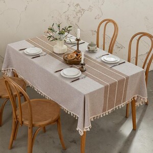 May include: A rectangular beige tablecloth with a brown and white striped center design. The tablecloth is set on a wooden table with wooden chairs. The table is set with white plates, silverware, and candles.