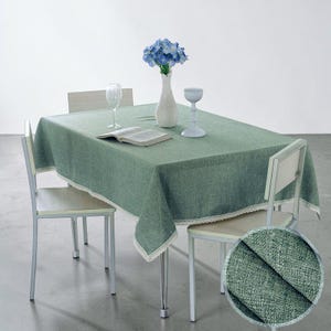 May include: A sage green rectangular tablecloth with a white lace trim, on a table with four chairs. A white vase with blue flowers, a wine glass, an open book, and a decorative candle holder are on the table.