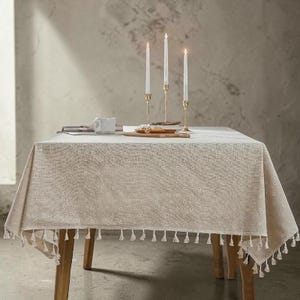 May include: A beige tablecloth with a repeating floral pattern and tassel trim, draped over a wooden table. Three white candles in gold holders and a small wooden serving board are on the table, creating a warm, inviting atmosphere.
