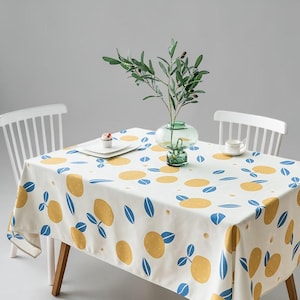 May include: A white tablecloth with a repeating pattern of yellow lemons and blue leaves. The tablecloth is on a wooden table with white chairs. A glass vase with green stems and a small glass vase are on the table.