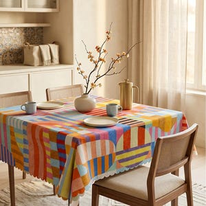 May include: A square tablecloth featuring a vibrant, multicolored geometric pattern with stripes, circles, and wavy lines in shades of orange, blue, yellow, and pink. The tablecloth has a scalloped edge and is draped over a wooden dining table.