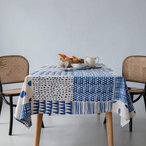 May include: A rectangular tablecloth with a blue and white geometric pattern covers a wooden table. The pattern includes dots, triangles, and lines. Two woven chairs flank the table, and a small meal is set on top.