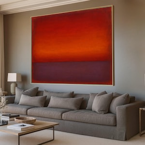 Puede incluir: Gran pintura abstracta con un degradado de tonos rojos, naranjas y morados, enmarcada en dorado. La obra cuelga sobre un sofá gris con varios cojines, creando una estética de salón moderno.