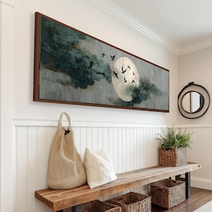 May include: A rectangular wall art piece with a dark brown frame. The artwork depicts a moon with birds flying in front of it, set against a backdrop of teal and gray clouds. The artwork is hanging on a white wall.