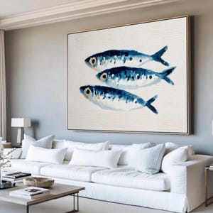 Könnte beinhalten: Gerahmtes Leinwandkunstwerk mit drei stilisierten blauen Fischen auf weißem Hintergrund. Die Fische haben dunkelblaue Markierungen und gelbe Augen. Das Kunstwerk wird in einem Wohnzimmer gezeigt.