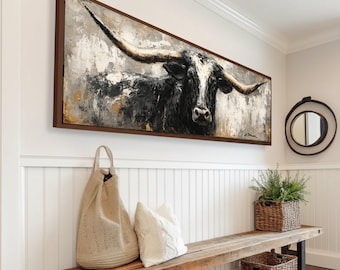 May include: A large rectangular framed painting of a longhorn bull with large horns. The artwork is primarily black, white, and gray with brown accents. The painting hangs above a wooden bench with baskets and a woven bag.