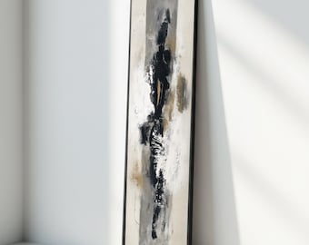 May include: Abstract art print in a black frame. The artwork features a vertical composition with a central black form, accented by gray, white, and gold brushstrokes. The piece leans against a white wall, showcasing a modern aesthetic.