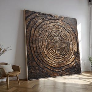 Puede incluir: Obra de arte grande y enmarcada que presenta una representación detallada y texturizada de anillos de árbol. La obra muestra círculos concéntricos en tonos marrones y negros, creando una estética natural. El marco es de color claro.