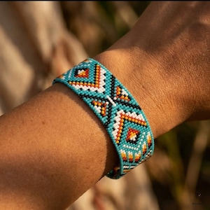 May include: A wide beaded bracelet featuring a geometric pattern in turquoise, white, black, orange, and yellow. The small, uniform beads are arranged in a repeating diamond and chevron design that wraps around the wrist, creating a textured surface.