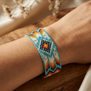 May include: A wide beaded bracelet featuring a geometric pattern in turquoise, white, orange, and yellow. The design consists of a central diamond shape surrounded by repeating chevron and triangular motifs, all crafted from small, uniform beads.