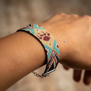 May include: A wide beaded bracelet featuring a geometric pattern in turquoise, beige, black, and white, with a central dark red bear paw print motif. The bracelet is finished with a silver-toned metal bar clasp and a small adjustable chain link.