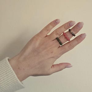 May include: Three beaded rings in shades of green, red, and brown are displayed on a hand. The rings are made of small, round beads. The hand is fair-skinned and the background is a plain, light beige color.