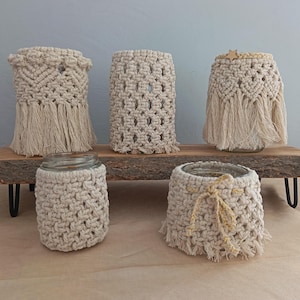 Peut inclure: Cinq cache-pots en macramé beige avec différents motifs et franges, présentés sur une étagère en bois avec des pieds en métal noir. Les pots sont vides, mettant en valeur les textures et les motifs tissés.