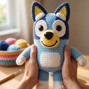 Könnte beinhalten: Handgefertigtes Bluey-Plüschtier aus Häkelarbeit, mit hellblauem Körper und dunkelblauen und gelben Akzenten. Das Spielzeug hat weiße Pfoten und einen lächelnden gelben Mund. Das Plüschtier wird vor einem Korb mit bunten Garnen gehalten.