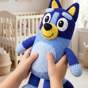May include: A handmade crochet plush toy in the shape of a cartoon dog. The toy features shades of blue, yellow, and white, with large eyes and a smiling face. The plush is held by a child's hands, showcasing its size and design.