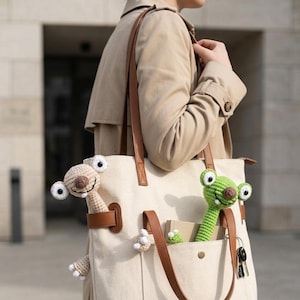 May include: A beige tote bag with brown leather straps and a front pocket. The pocket holds a green crocheted frog and a small notepad. A crocheted sloth is peeking out of the bag. The bag is carried by a person wearing a tan trench coat.