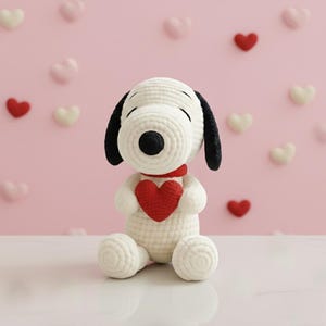 Puede incluir: Un peluche de Snoopy blanco hecho a mano con ganchillo, sosteniendo un corazón rojo. El peluche tiene orejas negras y una nariz negra, con una bufanda roja alrededor del cuello. El fondo es rosa con decoraciones en forma de corazón.
