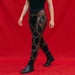 May include: A brown leather leg harness featuring a series of interconnected straps and metal rings worn over black leggings. The harness includes adjustable buckles and rivets, creating a cage-like structure that extends from the waist to the lower leg.