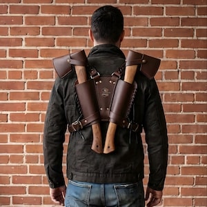 May include: A dark brown leather dual axe harness worn over a dark jacket. The harness features two holsters for axes with wooden handles and dark metal heads, secured with straps and buckles. The design includes lacing details and a central panel.