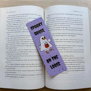 Purple bookmark with tiny black stars as the background with a goose dressed as a ghost with a candy bucket, with the words "spooky goose on the loose"