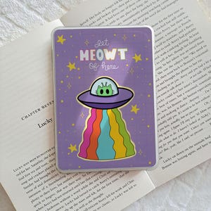 May include: A purple sticker featuring a cartoon cat in a UFO, with a rainbow beam and the text "Let MEOWT of here." The sticker has a gold border and star accents. A fun, whimsical design.