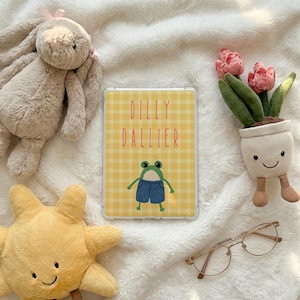 May include: A clear tablet case with a yellow gingham pattern and the words "DILLY DALLIER" in pink. A cartoon frog wearing blue overalls is on the case. The case is surrounded by plush toys, a potted plant, and gold-rimmed glasses.