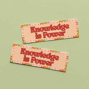 May include: Two rectangular bookmarks featuring a light peach background with the text Knowledge is Power in bold red letters. The borders are decorated with a repeating pattern of small orange, pink, and white flowers on a checkered background.