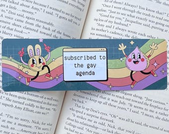 Pode incluir: Marcador de livros com um design retrô com um coelho de desenho animado e um personagem em forma de lágrima, com o texto "subscribed to the gay agenda" em uma tela de computador. O fundo tem listras de arco-íris.