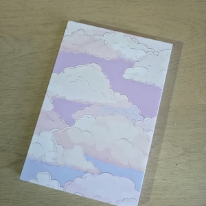 May include: A rectangular notebook featuring a dreamy cloudscape design. The cover showcases fluffy white clouds against a soft lavender and light blue sky. The notebook is positioned on a light brown wooden surface.