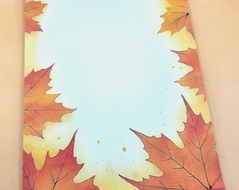 May include: A rectangular notepad with a border of orange and red autumn leaves. The leaves frame a light blue and white center. The notepad has a watercolor effect, with yellow and orange hues in the background.