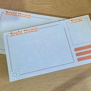 May include: Two light blue notepads with the words "Brain Dump" and "Notes" printed on them. The notepads have a dotted grid pattern and floral accents in orange and light blue. The notepads are on a wooden surface.