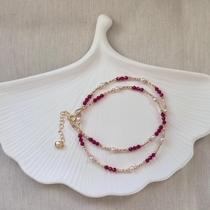 Può includere: Una delicata collana dorata a due fili. Un filo presenta piccole perline rotonde rosso scuro, l'altro ha piccole perline rotonde bianche. La collana è esposta su un piatto bianco a forma di foglia.